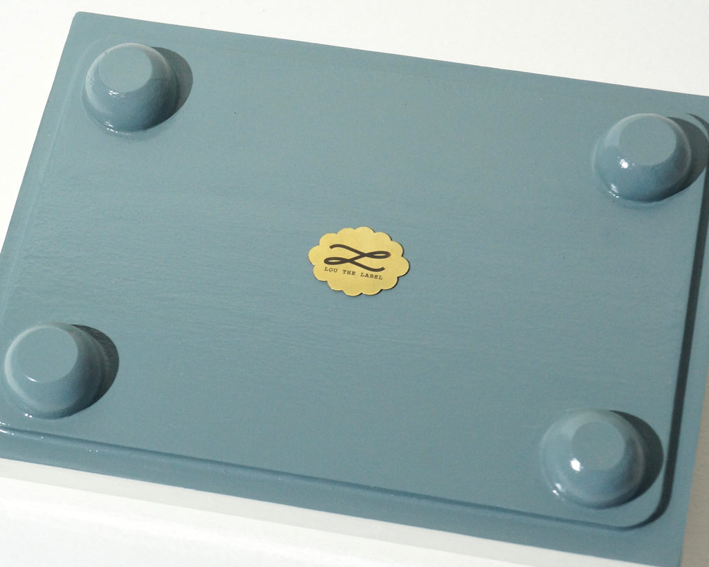 Close-up of the underside of a handmade jewelry tray showing the dusty blue painted finish, four rounded feet, and gold Lou The Label emblem.