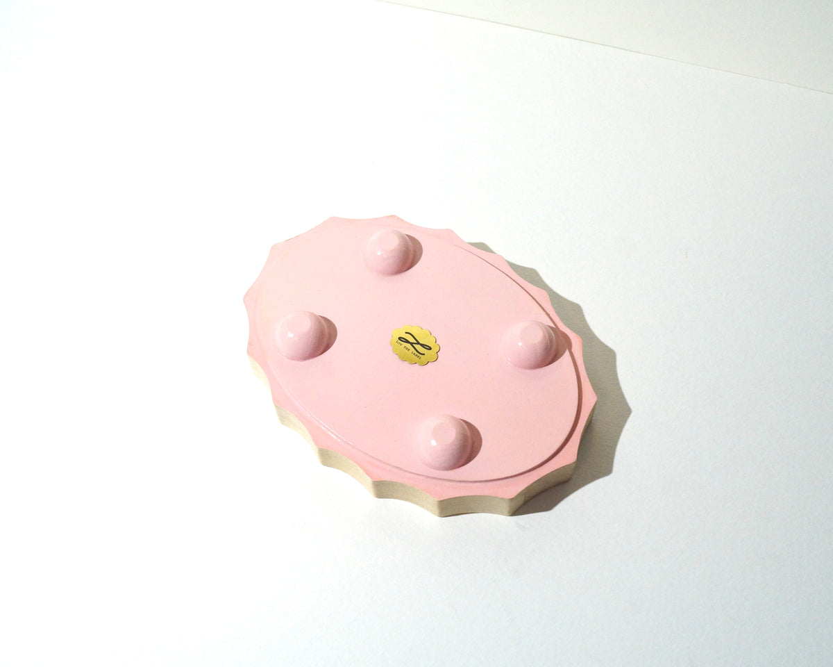 Bottom view of a Lou The Label handmade jewelry tray featuring a soft pink painted base, four rounded feet, natural wood edges, and a gold brand emblem in the center.