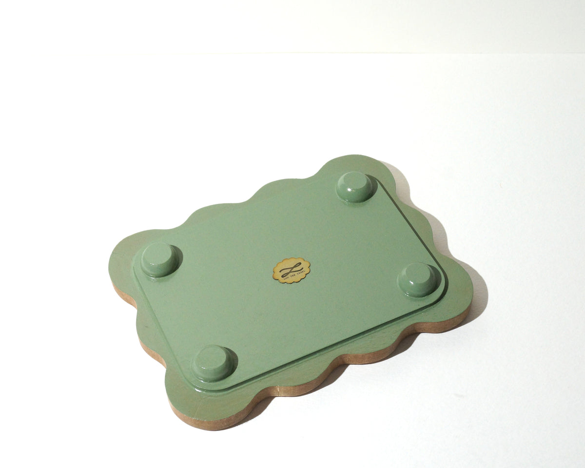 Bottom view of a handmade wooden jewelry tray with a sage green painted base, four rounded feet, and softly wavy edges, featuring a small gold Lou The Label brand emblem in the center.