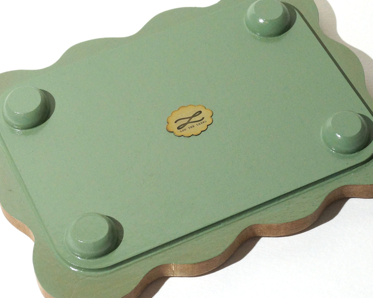 Bottom view of a handmade wooden jewelry tray with a sage green painted base, four rounded feet, and softly wavy edges, featuring a small gold Lou The Label brand emblem in the center.