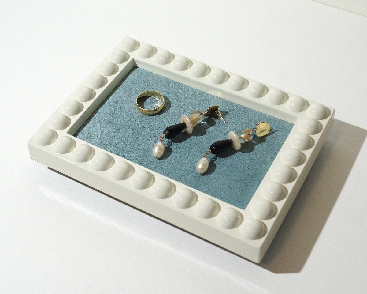 Lou The Label handmade rectangular jewelry tray featuring a glossy ivory frame with rounded bubble detailing and a dusty blue faux suede lining, displaying a gold ring and statement earrings.