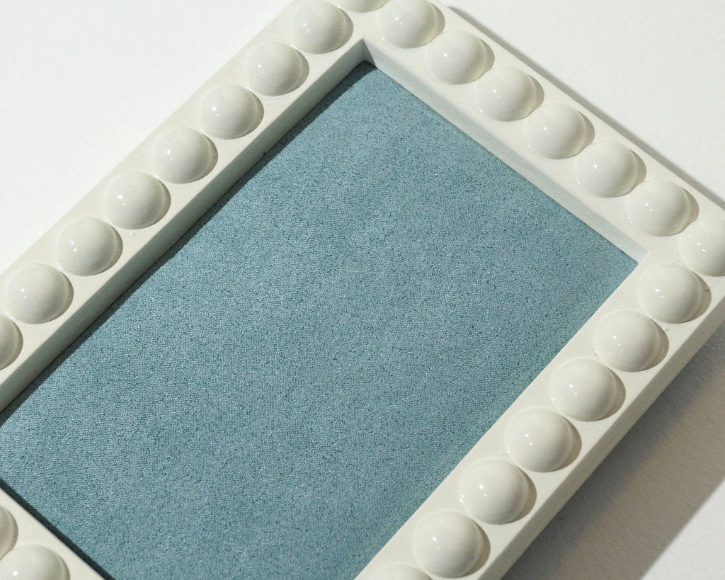 Close-up of a handmade jewelry tray highlighting the glossy ivory bubble-edged frame and textured dusty blue faux suede lining.