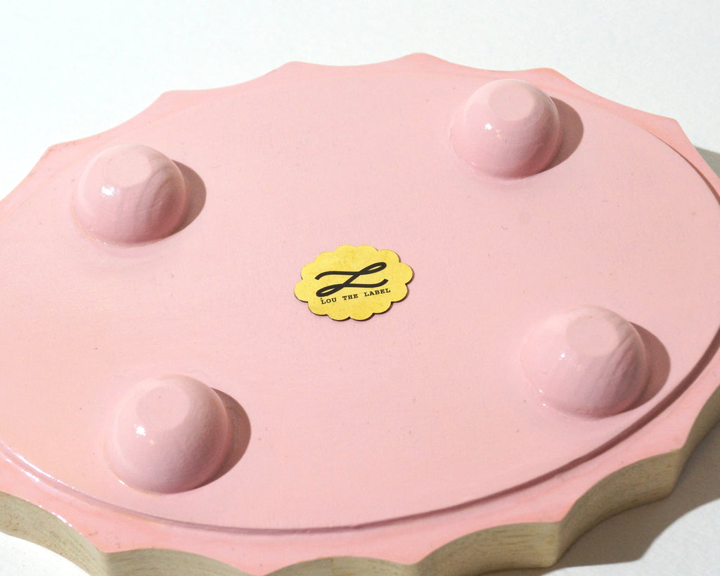 Close-up of the underside of a Lou The Label handmade oval jewelry tray featuring a soft pink painted base, four rounded feet, scalloped natural wood edges, and a gold brand emblem in the center.
