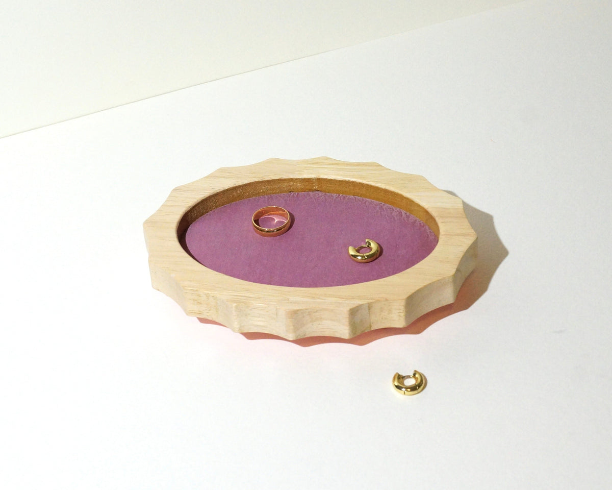 Angled view of a handmade oval wooden jewelry tray with scalloped edges and mauve pink brushed linen lining, displaying a gold ring and small gold hoop earrings.
