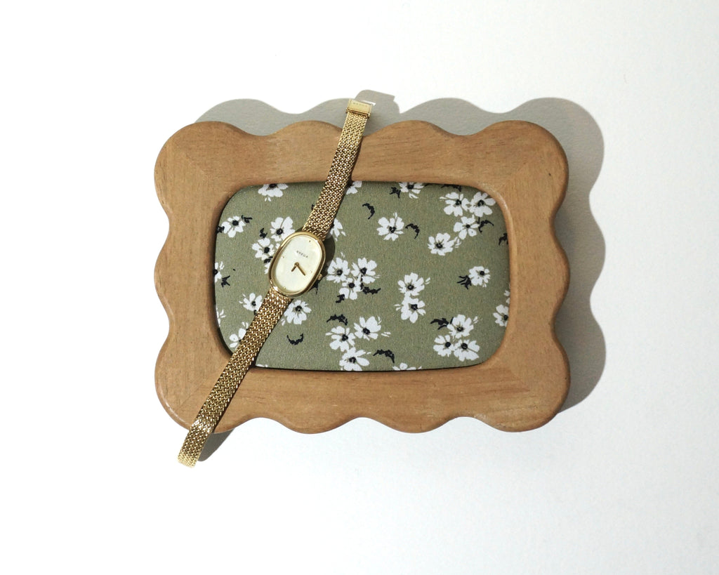 A handmade wooden jewelry tray with softly curved, wavy edges and sage green floral fabric lining, featuring a delicate gold wristwatch resting across the frame photographed on a white background. 