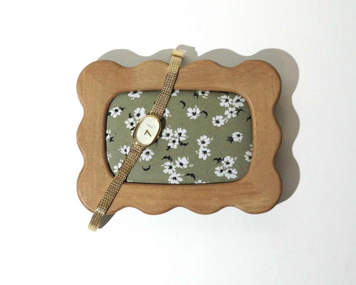 A handmade wooden jewelry tray with softly curved, wavy edges and sage green floral fabric lining, featuring a delicate gold wristwatch resting across the frame photographed on a white background. 