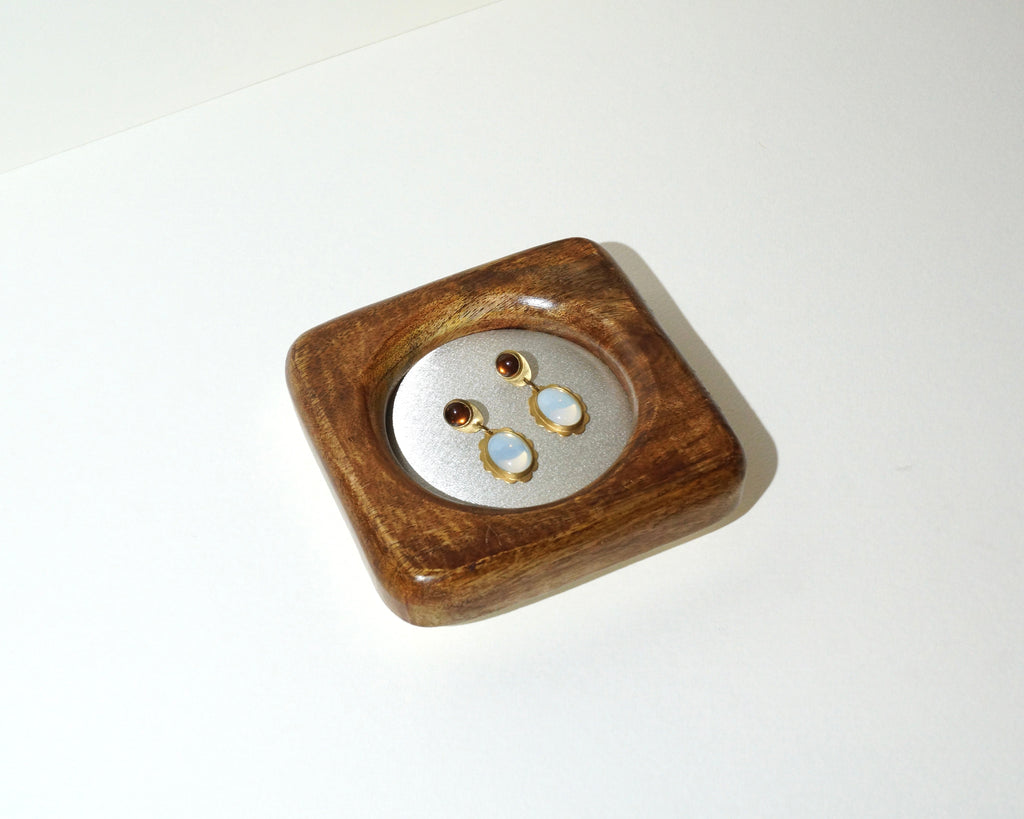 Overhead view of a handmade wooden jewelry tray with a circular ivory satin center, displaying a pair of gold drop earrings with pale blue stones and dark brown accents.
