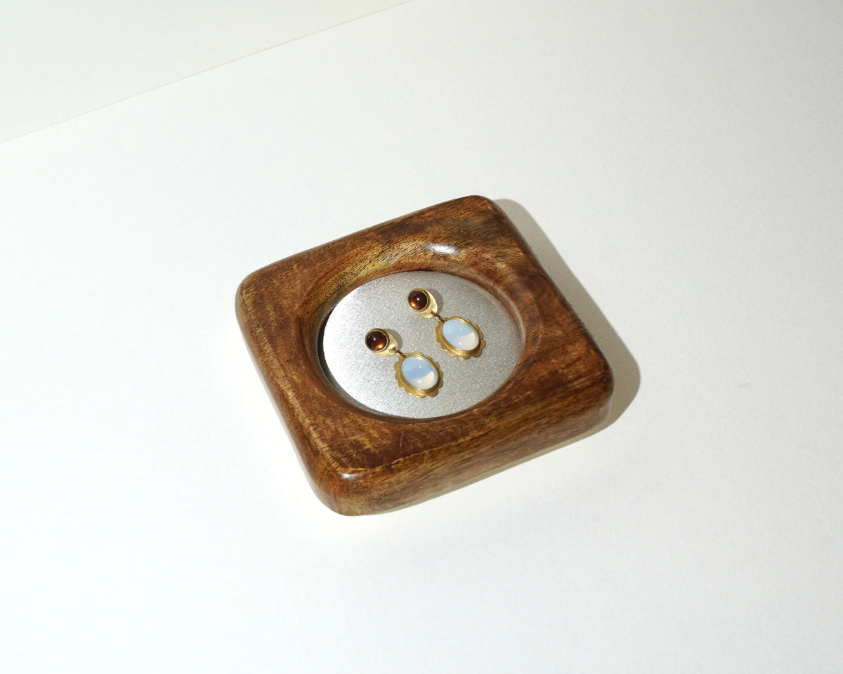 Overhead view of a handmade wooden jewelry tray with a circular ivory satin center, displaying a pair of gold drop earrings with pale blue stones and dark brown accents.