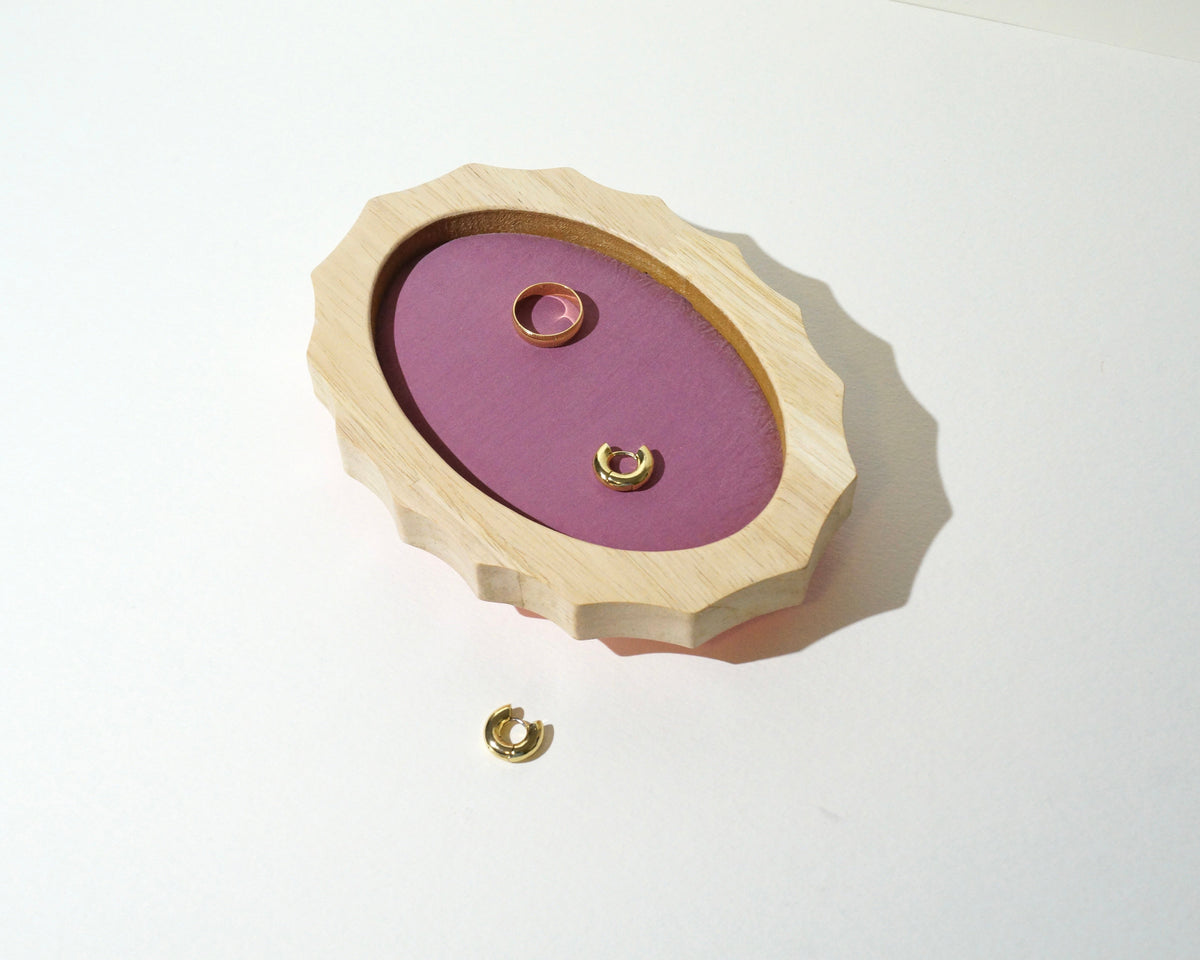 Overhead view of a handmade oval jewelry tray with a scalloped natural wood frame and mauve pink brushed linen lining, displaying gold rings and earrings.