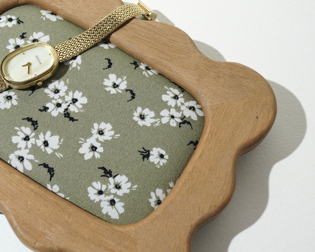 Close-up of a handmade wooden jewelry tray with softly curved, wavy edges and sage green floral fabric lining, featuring a delicate gold wristwatch resting across the frame photographed on a white background. 