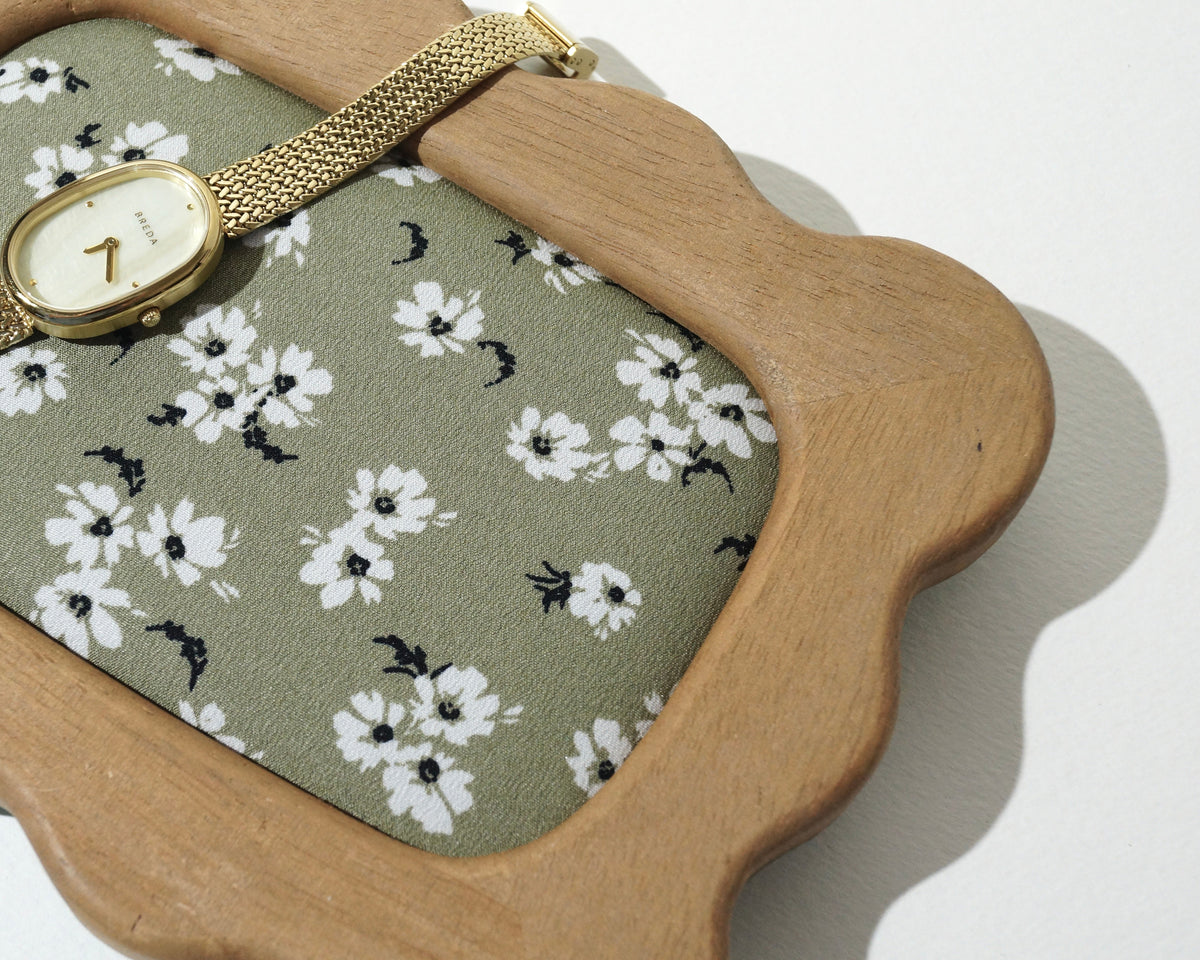 Close-up of a handmade wooden jewelry tray with softly curved, wavy edges and sage green floral fabric lining, featuring a delicate gold wristwatch resting across the frame photographed on a white background. 