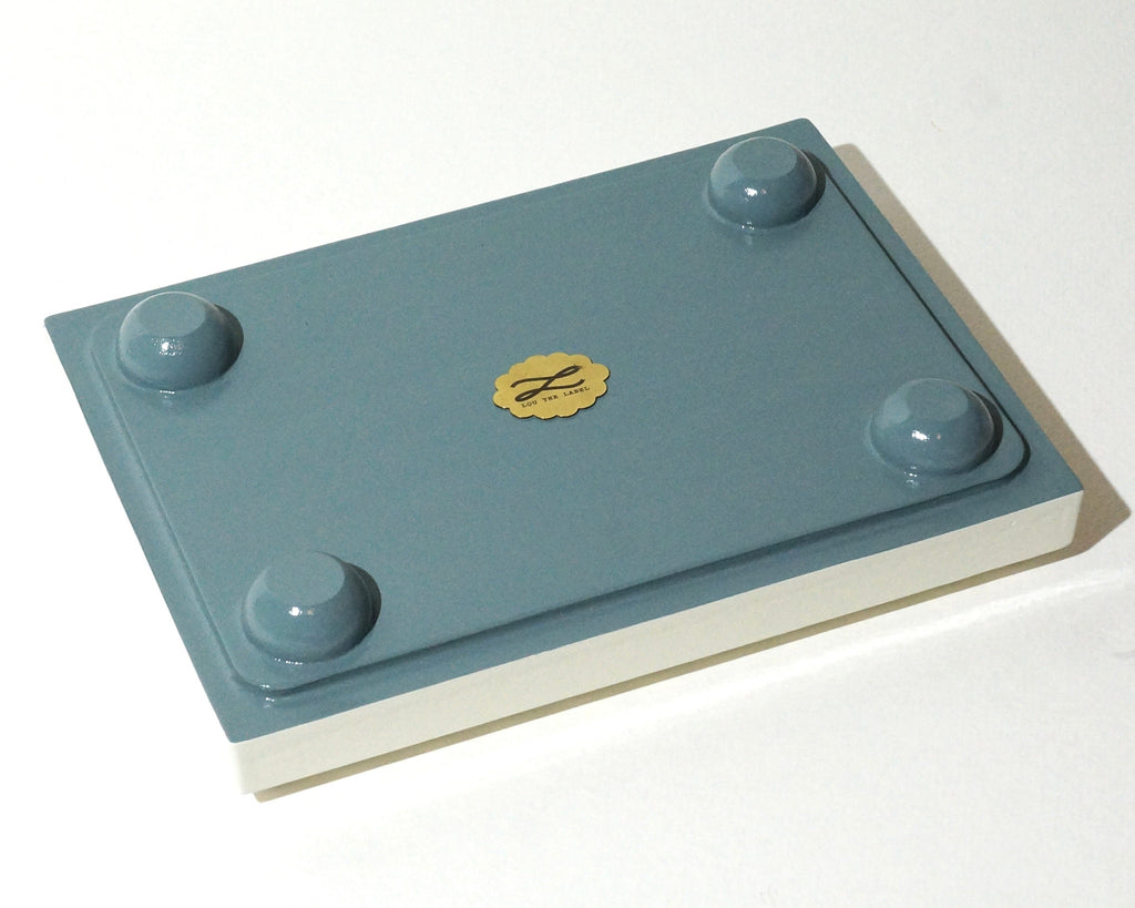 Bottom view of a Lou The Label jewelry tray featuring a dusty blue painted base, four rounded feet, and a gold brand emblem in the center.